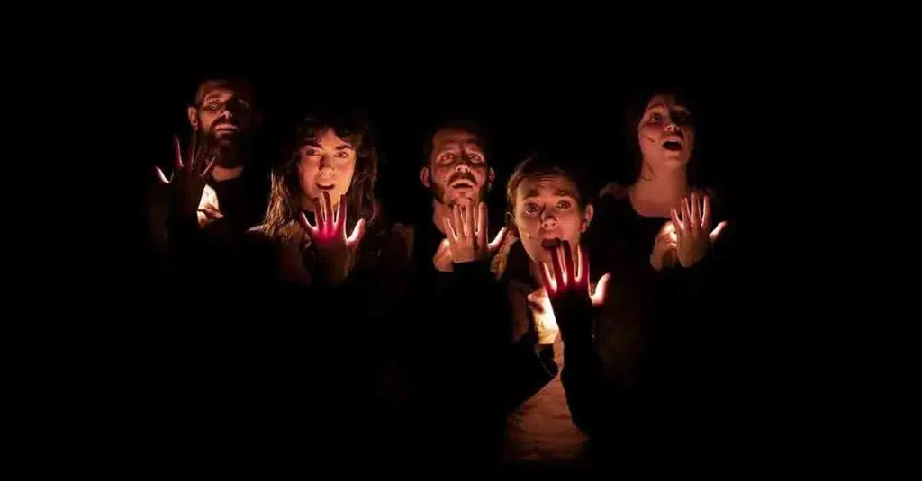 Cinque performer illuminano i volti e le mani con luci portatili nell’oscurità