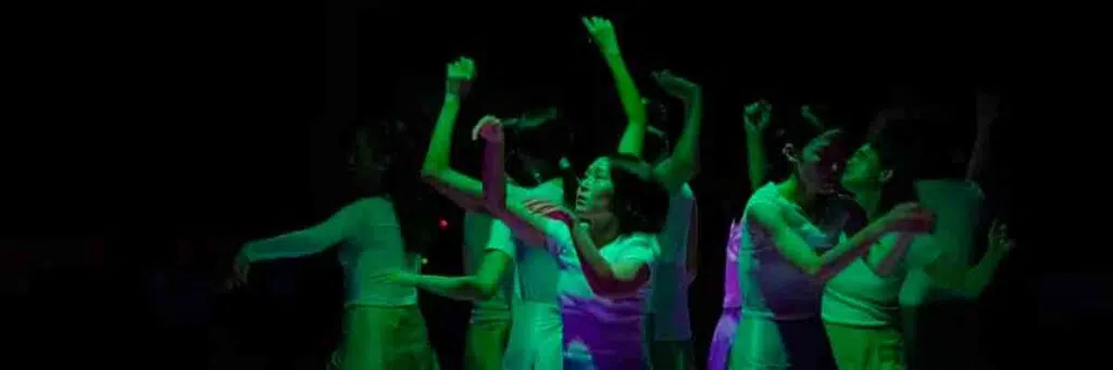 Ensemble di performer di Shimmering in scena sotto luci verdi e viola, con le braccia sollevate