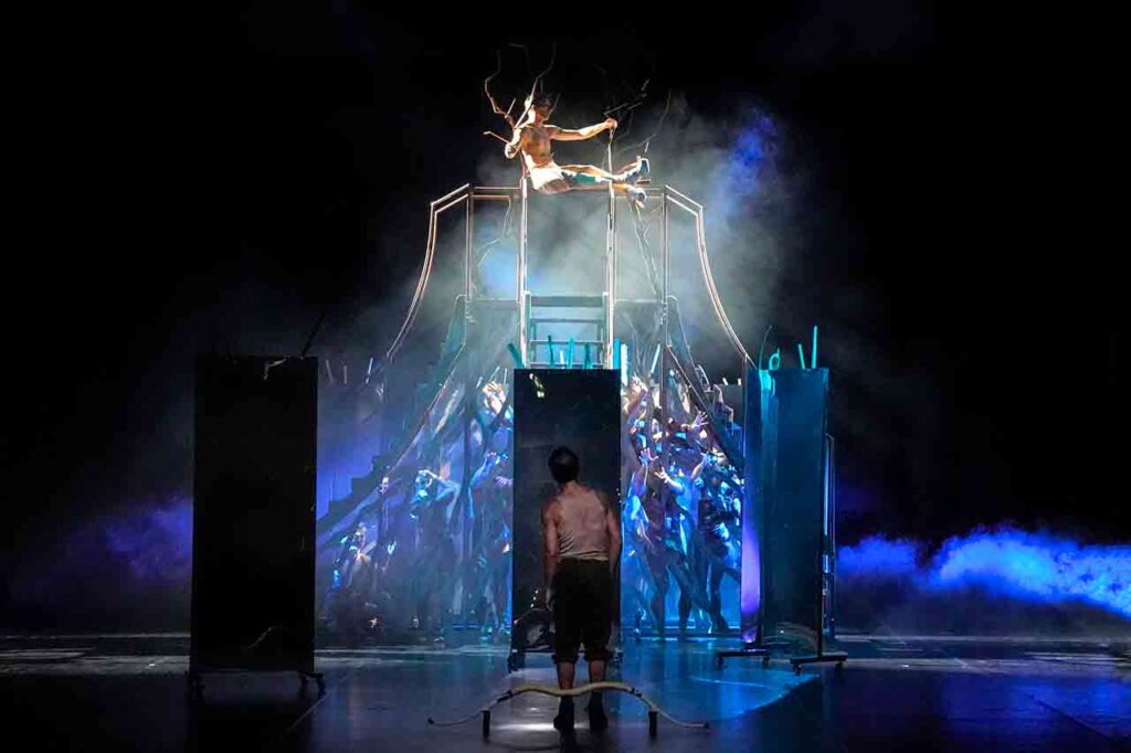 Danzatori si muovono sopra e dentro una struttura metallica avvolta da fumo e luci blu